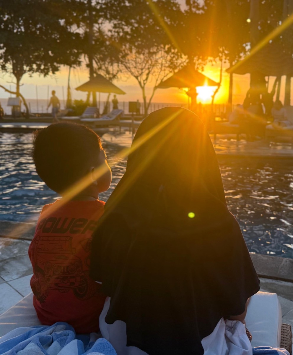 A woman in a headscarf and a child sitting by a pool at sunset, looking out over the sea on Lombok.