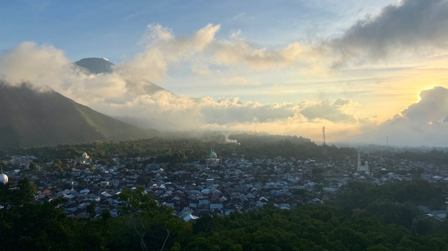 A Lombok village at sunrise, mosque domes among the rooftops and Mount Rinjani rising through the cloud behind it.