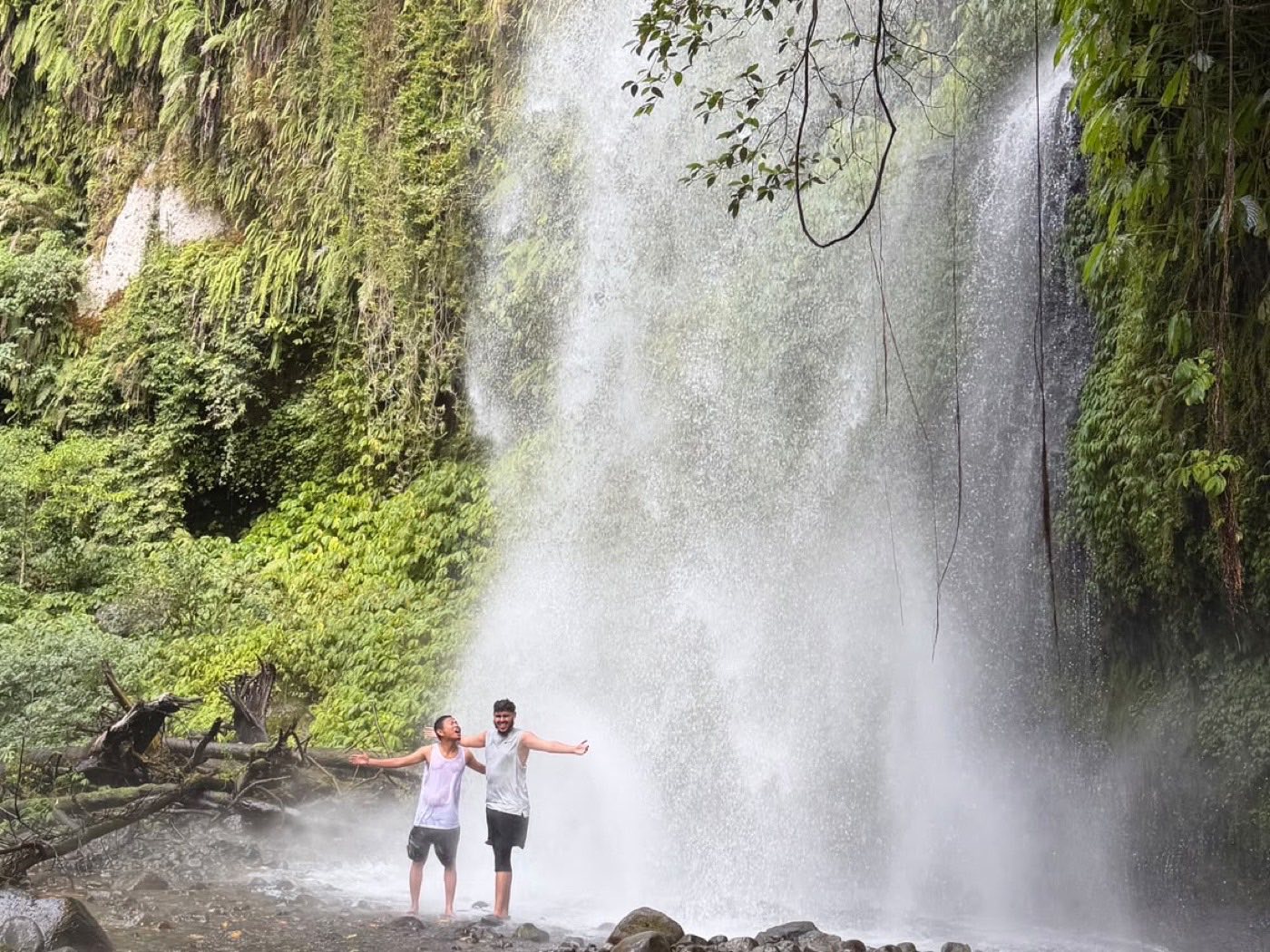 Two visitors standing arms out under a tall jungle waterfall in northern Lombok.