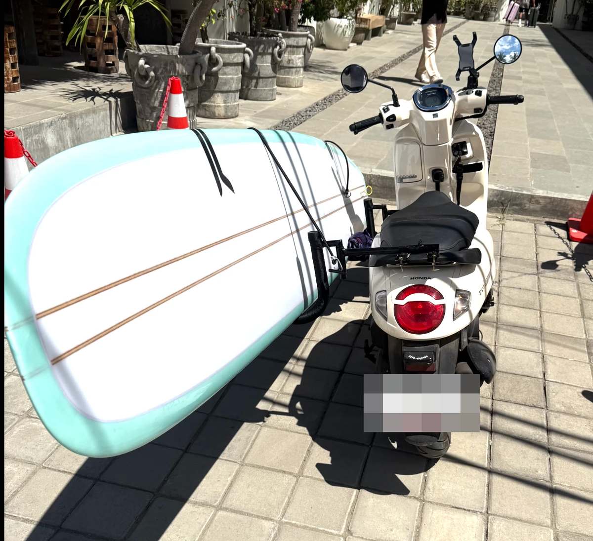 A rental scooter with a surfboard strapped to a side rack, parked on a paved street on Lombok.